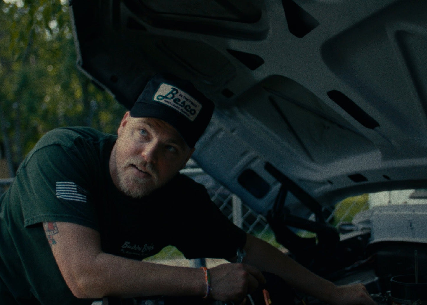 A man wearing a Besco “In The Pines” hat works under the hood of a classic truck during the Besco Release 1 shoot.