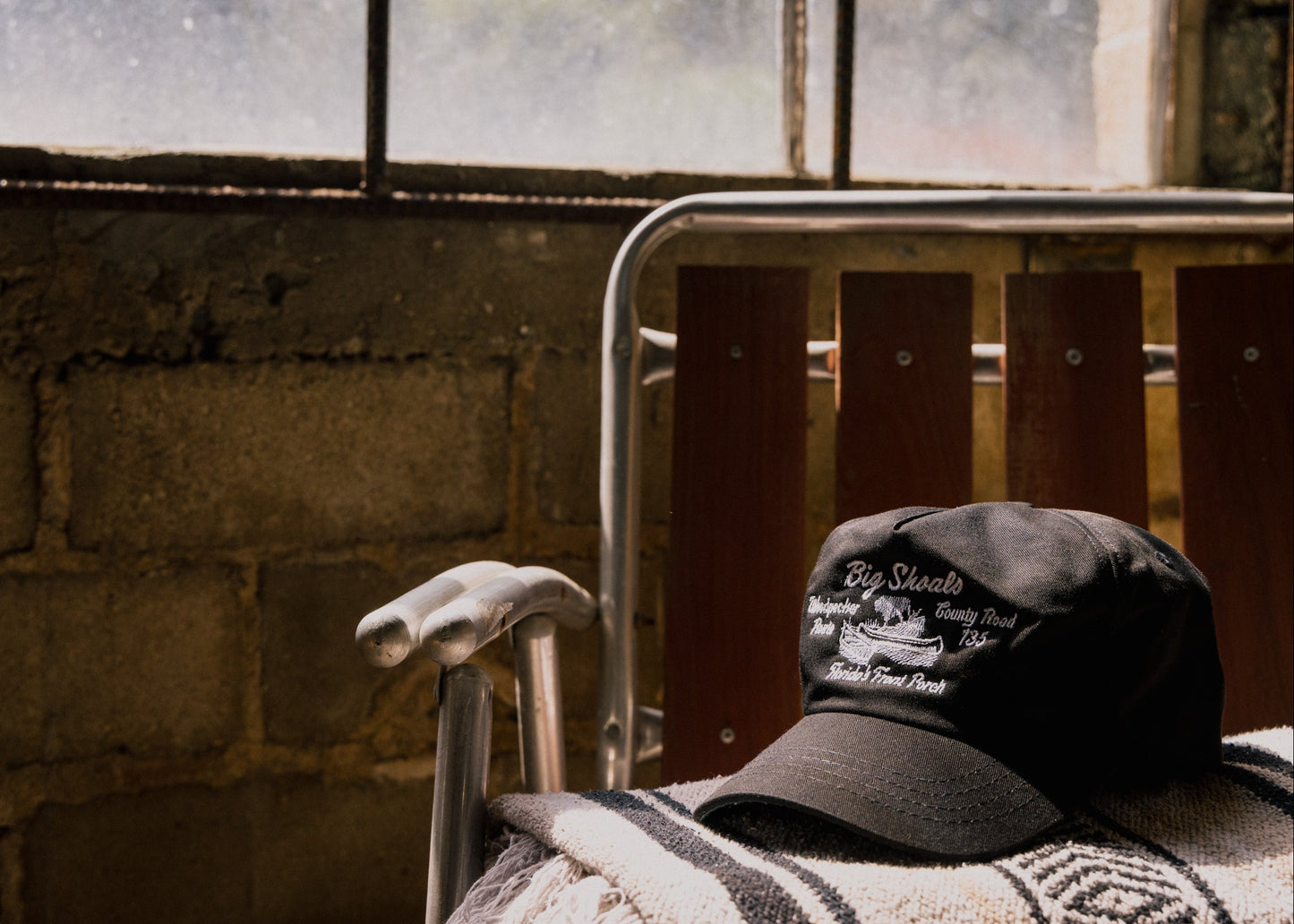 Black Besco ‘Big Shoals’ hat with white embroidery resting on a striped blanket draped over a vintage aluminum and wood folding chair, positioned in front of a rustic window with sunlight filtering through.