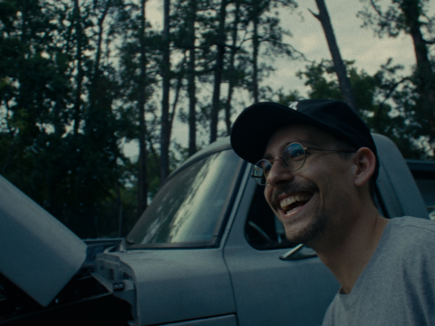 A man wearing a letter B Besco hat laughs beside a gray vintage truck with the hood open during the Besco Release 1 shoot.